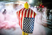 Protestujúci, zahalený v pozmenenej americkej vlajke, sleduje, ako sa poriadkové sily snažia rozohnať protestujúcich v Portlande.